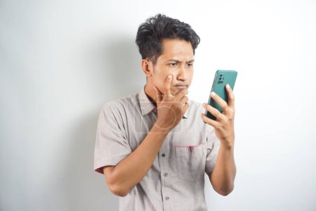 Photo for Thoughtful handsome asian man standing on white background with smartphone in hand, looking and thinking wearing shirt. Isolated. Copy space - Royalty Free Image