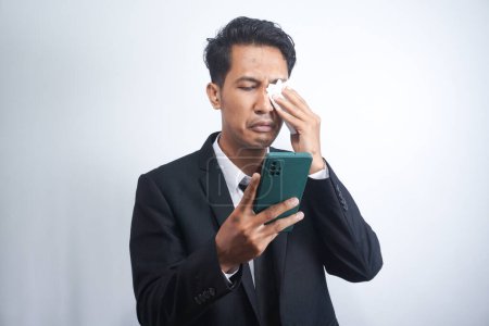 Photo for Asian guy wearing a suit looks sad reading online news from his cellphone. man shows disappointed gesture by wiping tears from his face - Royalty Free Image