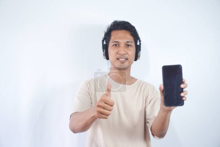 Photo for Half length shot of optimistic man holds smartphone with mock up screen, listens favourite soundtrack in headphones, wears casual cream t shirt, stands indoor. People and technology concept. - Royalty Free Image