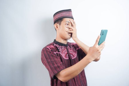 Photo for Amazed shocked asian guy holding smartphone in his hand, looking at the phone in surprise, stunned facial expression, stands on isolated white background - Royalty Free Image