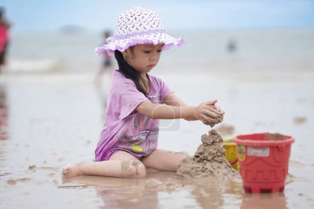 Photo for Balikpapan, March 1, 2023. A cute little girl wearing a purple beach hat and shirt is playing in the sand on the beach. Vacation and relax.Playful active kid on beach in summer vacation and child development. - Royalty Free Image