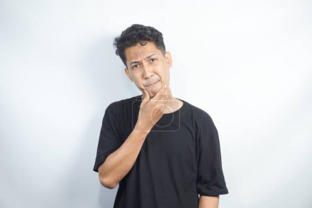 Photo for Portrait of unhappy determined Asian male  touching chin while thinking and looking with serious and worried look at camera, standing against white background. - Royalty Free Image