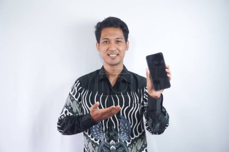 Photo for Shot of handsome guy in batik shirt isolated on grey background, presenting smart phone and pointing with finger at blank black screen with copyspace for ads. Expression of surprise and delight - Royalty Free Image