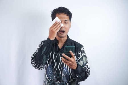 Photo for Young Asian man crying and sad while looking at smartphone. Crying and wiping tears with tissue, Indonesian man wearing batik shirt isolated gray background. - Royalty Free Image