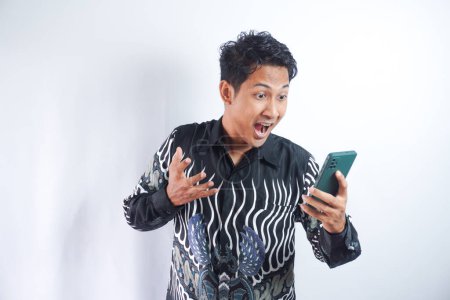 Photo for Young asian man with surprised facial expression hold smartphone in his hands, typing message, chatting with friends in social networks, playing online game, isolated against grey wall - Royalty Free Image