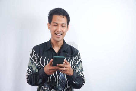 Photo for Image of smiling young man in batik shirt using mobile phone isolated by white background - Royalty Free Image