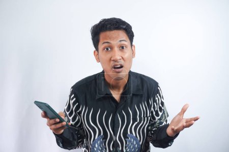 Téléchargez les photos : Beau asiatique homme portant chemise batik énervé en colère dans un geste furieux sur fond blanc - en image libre de droit
