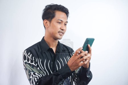 Photo for Portrait of a young cheerful excited asian man wearing batik shirt standing isolated over white background, using mobile phone - Royalty Free Image