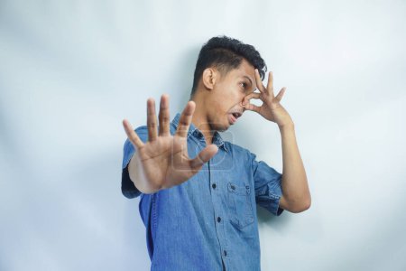 Photo for Stop unpleasant smell. Portrait of young man in denim casual shirt displeased by stink, grimacing in disgust and pinching his nose, making no gesture. indoor studio shot isolated on yellow background - Royalty Free Image