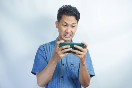 Foto de Joven hombre asiático jugando un videojuego en el teléfono móvil y obtuvo reacciones negativas. Las expresiones faciales pueden ser furiosas, feroces, molestas, infelices, irritadas, frustradas. - Imagen libre de derechos