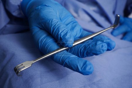 Doctor wearing blue gloves holding sharp Senn-Miller Retractor is a multi-purpose, dual-ended traction device that can be used to open superficial wounds in common surgical procedures