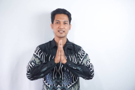 Photo for Smiling young Asian man wearing batik shirt, gesturing traditional greeting isolated over white background - Royalty Free Image