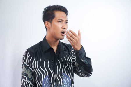 Téléchargez les photos : Mauvaise haleine. Concept d'halitose. Jeune homme portant une chemise batik vérifiant sa respiration avec sa main. - en image libre de droit
