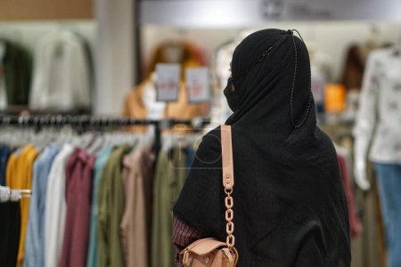 Photo for Beautiful young Asian woman in hijab looking at clothing products in shopping center. Attractive happy girl enjoying strolling in department store to choose new clothes and t-shirts from clothing racks in market center. - Royalty Free Image