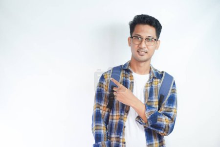 Photo for Cheerful Asian male student wearing casual clothes and backpack glasses pointing finger and palm at empty space isolated on white background. high school university college concept - Royalty Free Image