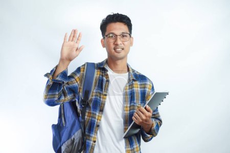 Asian teenager wearing student backpack and holding books waiving saying hello happy and smiling, friendly welcome gesture