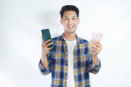 Adult Asian man smiling at the camera while holding phone and showing handful of money