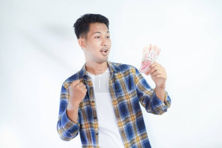 Téléchargez les photos : Adulte asiatique homme serré poing tout en tenant Indonésie papier monnaie et montrant expression excitée - en image libre de droit