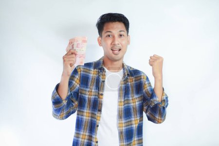 adulto asiático hombre apretado puño mientras celebración indonesia papel moneda y mostrando excitado expresión