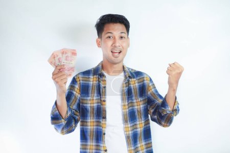 Foto de Adulto asiático hombre apretado puño mientras celebración indonesia papel moneda y mostrando excitado expresión - Imagen libre de derechos