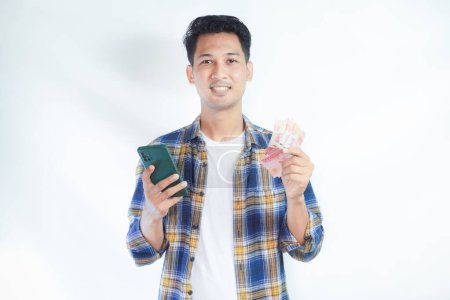 Foto de Adulto asiático hombre sonriendo a la cámara mientras sostiene el teléfono y mostrando un puñado de dinero - Imagen libre de derechos