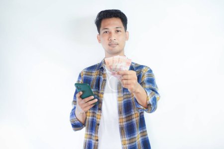 Photo for Adult Asian man smiling at the camera while holding phone and showing handful of money - Royalty Free Image