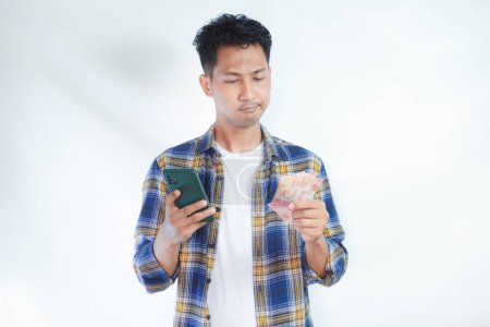 Erwachsener Asiate zeigt verwirrten Gesichtsausdruck, wenn er Handy und Papiergeld in der Hand hält