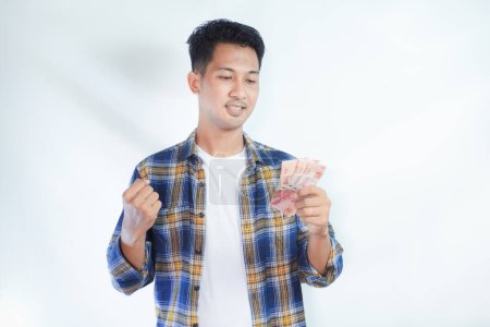 Téléchargez les photos : Adulte asiatique homme serré poing tout en tenant Indonésie papier monnaie et montrant expression excitée - en image libre de droit