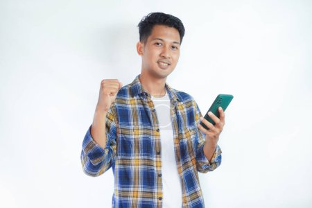 Photo for Adult Asian man showing excited expression while clenched fist and holding mobile phone - Royalty Free Image