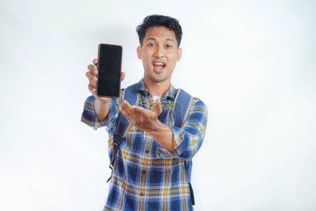 Photo for Cheerful young Asian man student in casual clothes and backpack glasses pointing finger at mobile phone with blank screen, recommending app isolated on white background. high school university college concept - Royalty Free Image