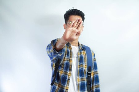 Photo for Dissatisfied young Asian student man in casual clothes with backpack showing a stop gesture with his palm, looking at the camera isolated on white background. High school university college concept - Royalty Free Image