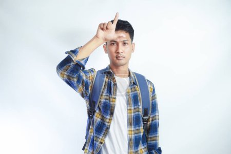 Unhappy Young Asian student giving loser sign on forehead, looking at you, disgust on face isolated on white wall. Negative human emotion facial expression. High school university college concept