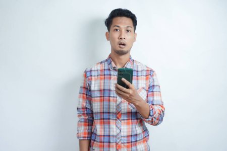 Hombre asiático adulto mirando a la cámara con expresión impactada mientras sostiene el teléfono