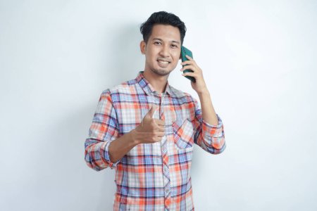 Photo for Adult Asian man wearing flannel shirt smiling and give thumb up while holding mobile phone - Royalty Free Image