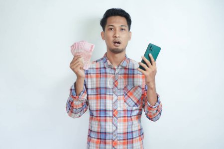 Hombre asiático adulto mostrando expresión de cara confusa al sostener el teléfono móvil y el papel moneda