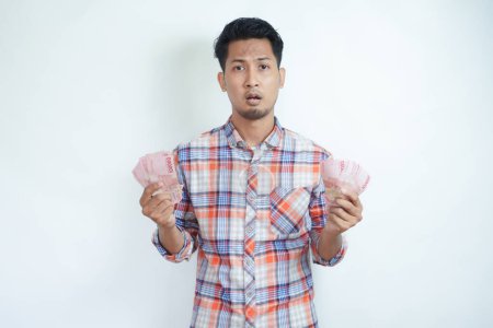 Adulto asiático hombre celebración de dinero y mirando cámara con expresión confusa
