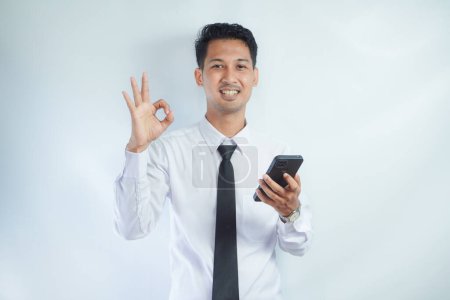 Erwachsener asiatischer Mann lächelt selbstbewusst und gibt OK Fingerzeichen, während er ein Mobiltelefon in der Hand hält