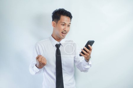 Photo for Adult Asian man clenching fist showing excitement when looking to his phone - Royalty Free Image