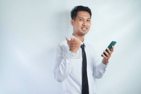 Photo for Adult Asian man wearing white shirt and tie smiling and give thumb up while holding mobile phone - Royalty Free Image