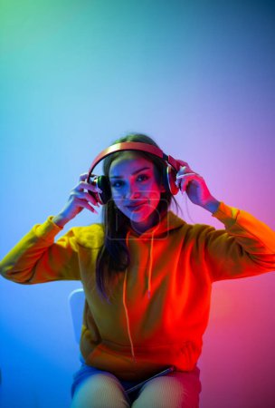 Photo for Shot of a young adult female wearing headphones, plying games, listening to music. Keeping in touch with others in a joyful and confident gesture. Modern studio with vibrant neon lighting. - Royalty Free Image