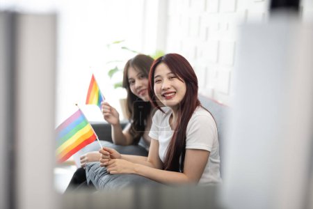 Foto de Un disparo de adorables parejas lesbianas asiáticas sostienen y ondean diminutas banderas LGBT mientras sonríen, se sienten confiados y orgullosos. Acciones vivas y armoniosas que muestren la igualdad de género en la sociedad - Imagen libre de derechos
