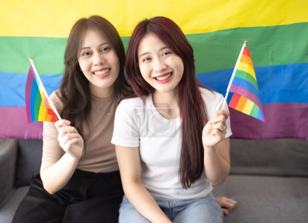 Téléchargez les photos : Tourné de beaux couples lesbiennes asiatiques tiennent et agitent de minuscules drapeaux LGBT tout en souriant, en étant confiant et en se sentant fier. Des actions à la fois vivantes et harmonieuses montrant l'égalité des sexes dans la société - en image libre de droit