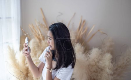 Téléchargez les photos : Une jeune fille asiatique avec bretelles applique le maquillage, regardant dans un miroir de poche dans une chambre confortable et bien éclairée. - en image libre de droit