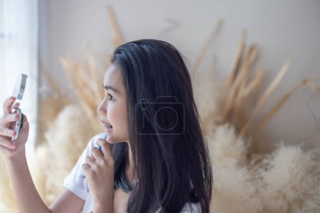 Foto de Una joven asiática con aparatos ortopédicos aplica maquillaje, mirando en un espejo de mano en una habitación acogedora y bien iluminada. - Imagen libre de derechos