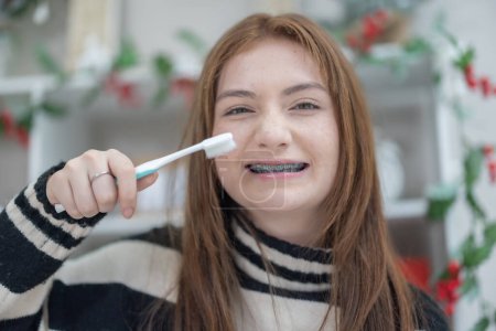 Foto de Una adolescente de raza mixta con frenos dentales sonríe mientras sostiene un cepillo de dientes en una habitación decorada festiva. - Imagen libre de derechos