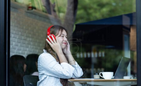Photo for A contemplative Asian woman listens to music on red headphones while working on her laptop in a cozy cafe setting. - Royalty Free Image
