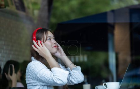 Foto de Una mujer asiática contemplativa escucha música en auriculares rojos mientras trabaja en su computadora portátil en una acogedora cafetería. - Imagen libre de derechos
