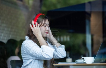 Foto de Una mujer asiática contemplativa escucha música en auriculares rojos mientras trabaja en su computadora portátil en una acogedora cafetería. - Imagen libre de derechos
