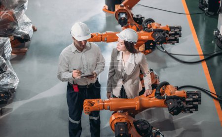 Foto de Ingeniero superior proporciona orientación a un aprendiz en medio de brazos robóticos en un entorno de fábrica de almacén de alta tecnología. - Imagen libre de derechos