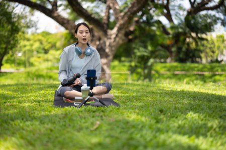 Photo for A female social media influencer is seated on a mat in a lush park, showcasing how to use a massage gun while recording the demonstration for her audience. - Royalty Free Image